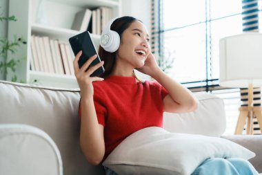 Genç Asyalı kadın evdeki kanepede dinleniyor, kulaklıkla cep telefonuyla müzik dinliyor. Halk yaşam tarzı konsepti