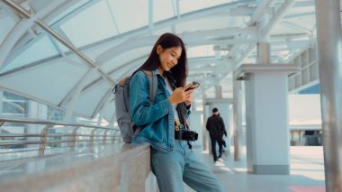 Dışarıda cep telefonu kullanan Asyalı genç bir kadın gülümsüyor. Kot ceket giyen ve halka açık bir yerde akıllı telefon tutan mutlu kadın turist.