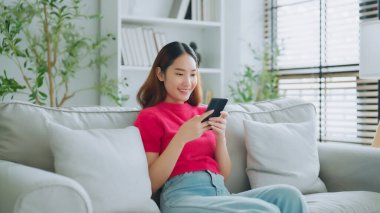 Mutlu genç Asyalı kadın rahat bir kanepede rahat rahat oturup akıllı telefonuna mesaj atıyor, gülümseyen kız cep telefonu sohbetini kullanıyor, evde online alışveriş yapıyor, video bağlantısı kuruyor.