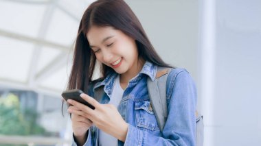 Dışarıda cep telefonu kullanan Asyalı genç bir kadın gülümsüyor. Kot ceket giyen ve halka açık bir yerde akıllı telefon tutan mutlu kadın turist.