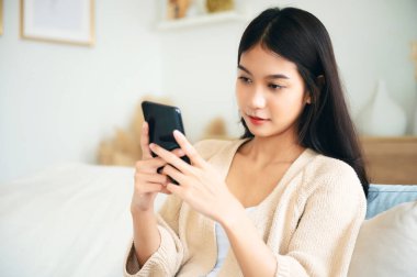 Happy young asian woman relax on comfortable couch at home texting messaging on smartphone, smiling girl use cellphone chatting, browse wireless internet on gadget, shopping online from home