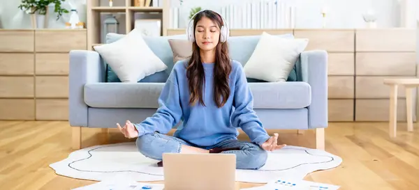 Happy young Asian woman practicing yoga and meditation at home sitting on floor in living room in lotus position and relaxing with closed eyes. Mindful meditation and wellbeing concept