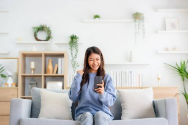 Happy young asian woman relax on comfortable couch at home texting messaging on smartphone, smiling girl use cellphone, chatting online message, shopping online from home
