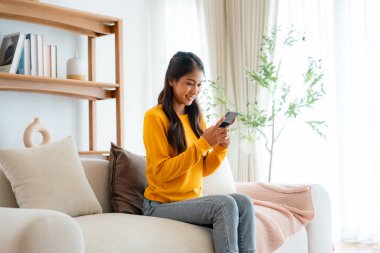 Mutlu genç Asyalı kadın rahat bir kanepede rahat rahat oturup akıllı telefonuna mesaj atıyor, gülümseyen kız cep telefonu sohbetini kullanıyor, cihazda kablosuz interneti tarıyor, evden online alışveriş yapıyor.