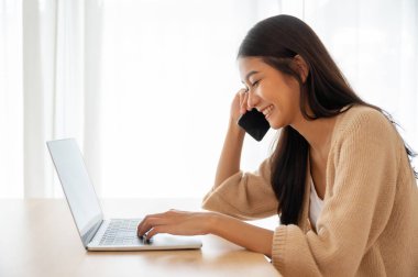 Genç Asyalı kadın evdeki oturma odasındaki masada bilgisayarla çalışırken cep telefonuyla konuşuyor.