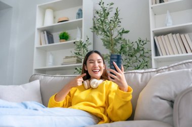 Mutlu Asyalı genç kadın kulaklık takıyor ve evdeki rahat koltukta dinleniyor, akıllı telefondan mesajlaşıyor, gülümseyen kız cep telefonu sohbetini kullanıyor, internetten alışveriş yapıyor, video görüşmesi yapıyor.