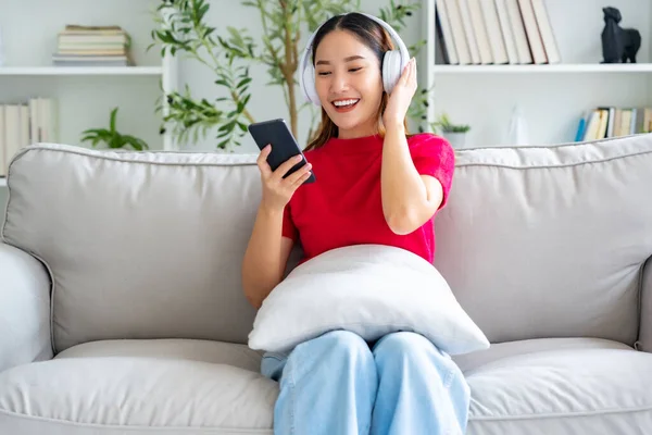 Young Asian woman relaxing on sofa at home, listening music with headset on cell phone. People lifestyle concept