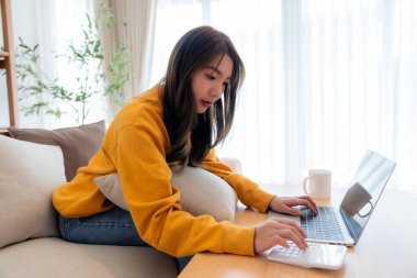 Genç Asyalı kadın gözlük takıyor dizüstü bilgisayar kullanıyor, oturma odasında çalışıyor, kahve fincanı masada. Konforlu ofis işyeri, uzak çalışma