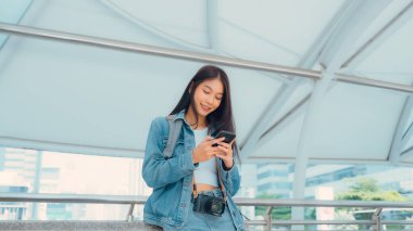 Dışarıda cep telefonu kullanan Asyalı genç bir kadın gülümsüyor. Kot ceket giyen ve halka açık bir yerde akıllı telefon tutan mutlu kadın turist.