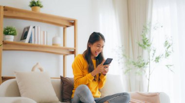 Happy young asian woman relax on comfortable couch at home texting messaging on smartphone, smiling girl use cellphone, chatting online message, shopping online from home
