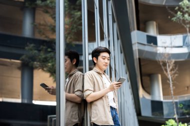 Akıllı telefon kullanan yakışıklı Asyalı öğrencinin portresi. Dışarıda duran genç bir adam elinde cep telefonuyla gülümsüyor.