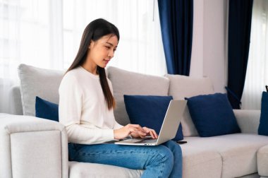 Genç Asyalı kadın evdeki koltukta otururken dizüstü bilgisayar kullanıyor.