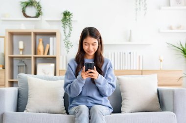 Happy young asian woman relax on comfortable couch at home texting messaging on smartphone, smiling girl use cellphone, chatting online message, shopping online from home
