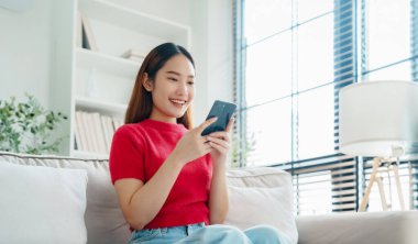 Mutlu genç Asyalı kadın rahat bir kanepede rahat rahat oturup akıllı telefonuna mesaj atıyor, gülümseyen kız cep telefonu sohbetini kullanıyor, evde online alışveriş yapıyor, video bağlantısı kuruyor.
