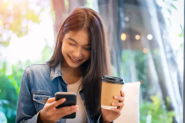 Asian woman happy and typing messages on smartphone in cafe. Smiling young woman sitting at table with hot cup of coffee and using mobile phone