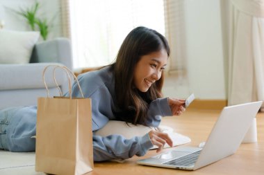 Genç Asyalı mutlu kadın evdeki oturma odasında bilgisayarla gülümsüyor, evde yerde yatıyor, banka kartından internet üzerinden ödeme yapıyor. Kredi kartıyla online alışveriş
