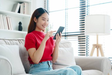 Mutlu genç Asyalı kadın rahat bir kanepede rahat rahat oturup akıllı telefonuna mesaj atıyor, gülümseyen kız cep telefonu sohbetini kullanıyor, evde online alışveriş yapıyor, video bağlantısı kuruyor.