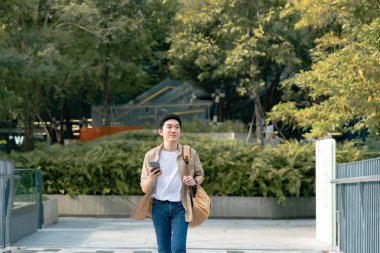 Akıllı telefon kullanan yakışıklı Asyalı öğrencinin portresi. Genç bir adam elinde cep telefonuyla mutlu bir şekilde dışarıda yürüyor.