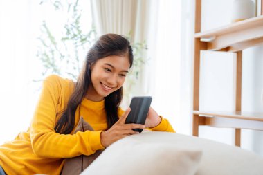 Cep telefonu teknolojisiyle kanepede oturan mutlu genç Asyalı kadın ecommerce alışverişi yapıyor, internetten alışveriş yapıyor evdeki rahat oturma odasındaki kanepede rahatlatıcı mesajlar yazıyor.