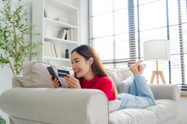 Oturma odasındaki kanepede uzanan Asyalı genç bir kadın internet üzerinden cep telefonundan banka ödemeleri yapıyor. Kredi kartıyla cep telefonundan alışveriş yapmak