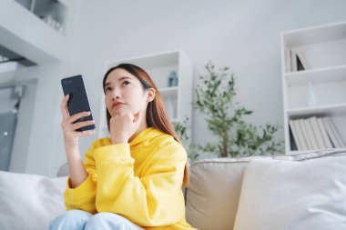 Mutlu genç Asyalı kadın rahat bir kanepede rahat rahat oturup akıllı telefonuna mesaj atıyor, gülümseyen kız cep telefonu sohbetini kullanıyor, evde online alışveriş yapıyor, video bağlantısı kuruyor.