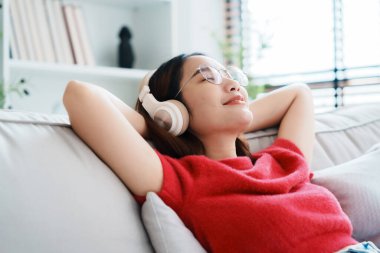 Rahat genç Asyalı kadın rahat koltukta dinleniyor, sakin çekici kız kanepede dinleniyor, temiz hava soluyor gözleri kapalı, gözlük takıyor ve kulaklık takıyor, evde meditasyon yapıyor.