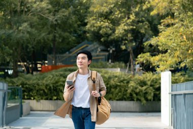 Akıllı telefon kullanan yakışıklı Asyalı öğrencinin portresi. Genç bir adam elinde cep telefonuyla mutlu bir şekilde dışarıda yürüyor.