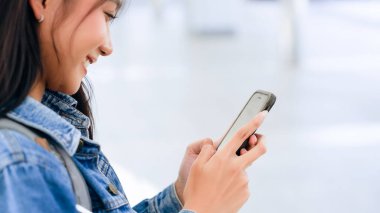 Dışarıda cep telefonu kullanan Asyalı genç bir kadın gülümsüyor. Kot ceket giyen ve halka açık bir yerde akıllı telefon tutan mutlu kadın turist.