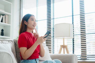 Mutlu genç Asyalı kadın rahat bir kanepede rahat rahat oturup akıllı telefonuna mesaj atıyor, gülümseyen kız cep telefonu sohbetini kullanıyor, evde online alışveriş yapıyor, video bağlantısı kuruyor.