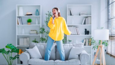 Oturma odasındaki kanepede genç bir Asyalı kadın dans ediyor. Gülümseyen Asyalı bir kadın, mutlu, zihinsel ve fiziksel olarak sağlıklı, kulaklık takıyor ve müzik dinliyor.