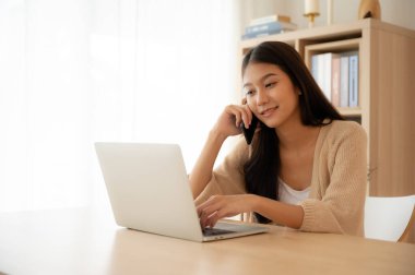 Genç Asyalı kadın evdeki oturma odasındaki masada bilgisayarla çalışırken cep telefonuyla konuşuyor.