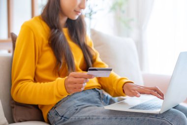 Oturma odasındaki kanepede oturan Asyalı genç bir kadın, internet üzerinden bilgisayar bilgisayarındaki banka kartından online bankacılık ödemeleri yapıyor. Kredi kartıyla internet üzerinden alışveriş yapmak