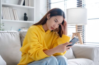 Bad news on device screen. Upset asian woman frustrated by problem with work or relationships, sitting on couch, feeling despair and anxiety, loneliness, having psychological trouble clipart