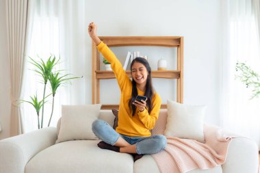 Happy young asian woman relax on comfortable couch at home texting messaging on smartphone, smiling girl use cellphone, chatting online message, shopping online from home