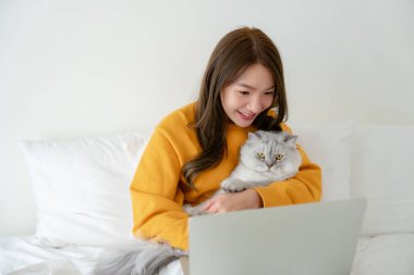 Evde çalışan Asyalı genç bir kadın yatak odasında kedicilik oynarken dizüstü bilgisayar kullanıyor.
