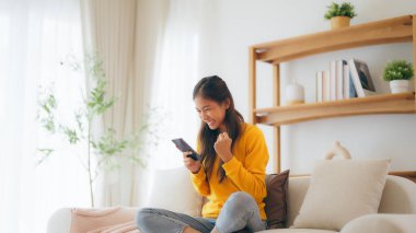Happy young asian woman relax on comfortable couch at home texting messaging on smartphone, smiling girl use cellphone, chatting online message, shopping online from home