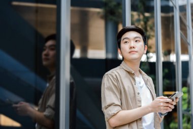 Akıllı telefon kullanan yakışıklı Asyalı öğrencinin portresi. Dışarıda duran genç bir adam elinde cep telefonuyla gülümsüyor.