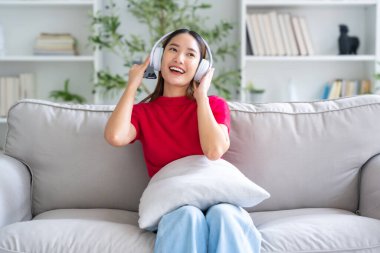 Genç Asyalı kadın evdeki kanepede dinleniyor, kulaklıkla cep telefonuyla müzik dinliyor. Halk yaşam tarzı konsepti