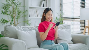 Mutlu genç Asyalı kadın rahat bir kanepede rahat rahat oturup akıllı telefonuna mesaj atıyor, gülümseyen kız cep telefonu sohbetini kullanıyor, evde online alışveriş yapıyor, video bağlantısı kuruyor.