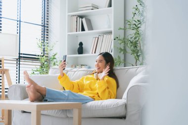 Mutlu Asyalı genç kadın kulaklık takıyor ve evdeki rahat koltukta dinleniyor, akıllı telefondan mesajlaşıyor, gülümseyen kız cep telefonu sohbetini kullanıyor, internetten alışveriş yapıyor, video görüşmesi yapıyor.