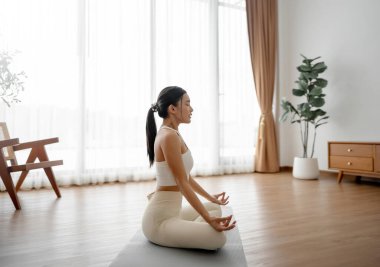 Genç Asyalı kadın evde nilüfer pozisyonunda meditasyon yapıyor, spor kıyafetleriyle yerde oturuyor. Rahatlama, huzur ve farkındalık için nefes alma egzersizi.
