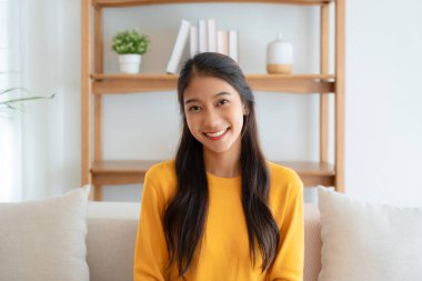 Oturma odasında kanepede oturmuş gülümseyen Asyalı genç kadının portresi ve kameraya bakmak mutlu yüz ifadesi, güzel cilt, sağlıklı bir vücut.