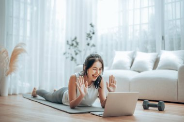 Genç Asyalı bir kadın laptopun önünde yoga minderinin üzerinde yerde yatıyor ve evde antrenman yapıyor. Egzersiz yapmadan önce internetten antrenörle iletişim kuran güzel, gülümseyen bir kadın.