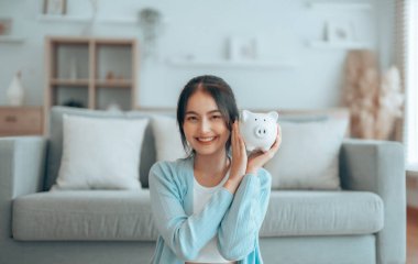 Genç Asyalı kadın domuz kumbarasını tutuyor ve evde yerde oturuyor. Para ve mali yatırım tasarrufu
