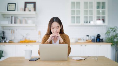 Asyalı genç bir kadın ciddi ciddi evdeki bilgisayar dizüstü bilgisayarında çalışıyor. Çözüm bulma sorununu düşünüyor.