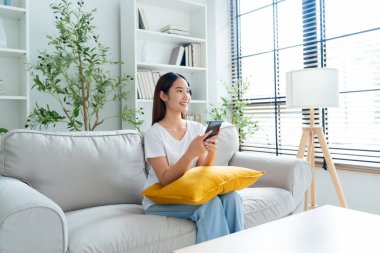 Mutlu genç Asyalı kadın rahat bir kanepede rahat rahat oturup akıllı telefonuna mesaj atıyor, gülümseyen kız cep telefonu sohbetini kullanıyor, evde online alışveriş yapıyor, video bağlantısı kuruyor.