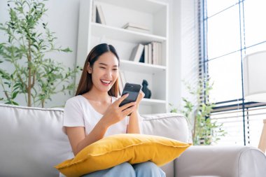 Happy young asian woman relax on comfortable couch at home texting messaging on smartphone, smiling girl use cellphone, chatting online message, shopping online from home
