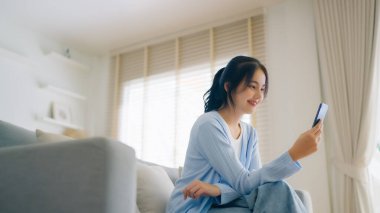 Oturma odasında komik mobil uygulamalar kullanarak rahat bir koltukta oturan gülümseyen genç Asyalı kadın. Evdeki Kadın, Çevrimiçi Alışveriş Yapıyor, Arkadaşlarına Mesajlaşıyor, Sosyal Medya 'ya Gönderiyor