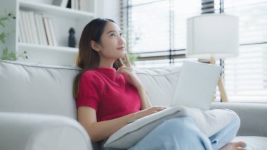 Evdeki kanepede otururken dizüstü bilgisayar kullanan mutlu genç Asyalı kadın.