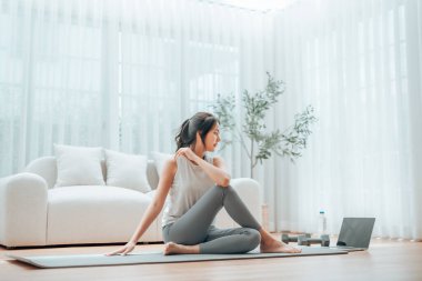 Genç Asyalı kadın oturma odasında bilgisayarlı dizüstü bilgisayarla antrenman yaparken yoga antrenmanını spor minderinde yapıyor.